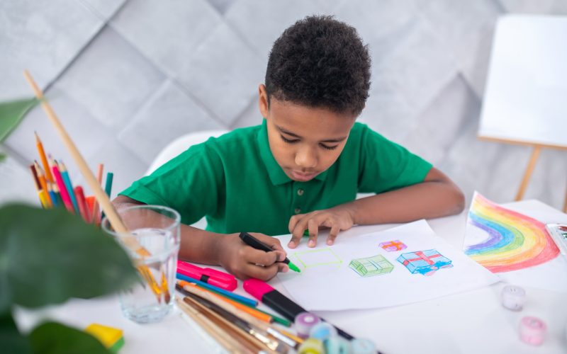 Drawing. Concentrated serious dark-skinned school-age boy drawing diligently with colored marker on sheet of paper sitting at table with art supplies in bright room